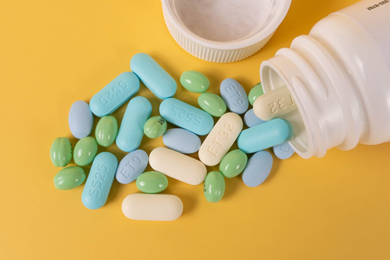 Various colored pills scattered from a white bottle on a yellow surface.