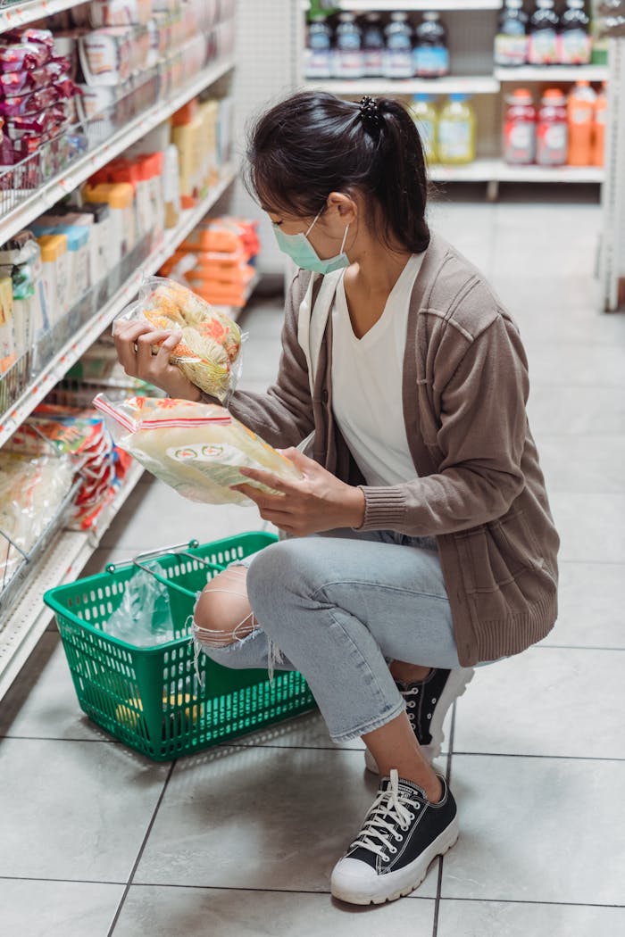 A woman in casual attire and face mask choosing groceries in a store aisle, emphasizing safety and protection.